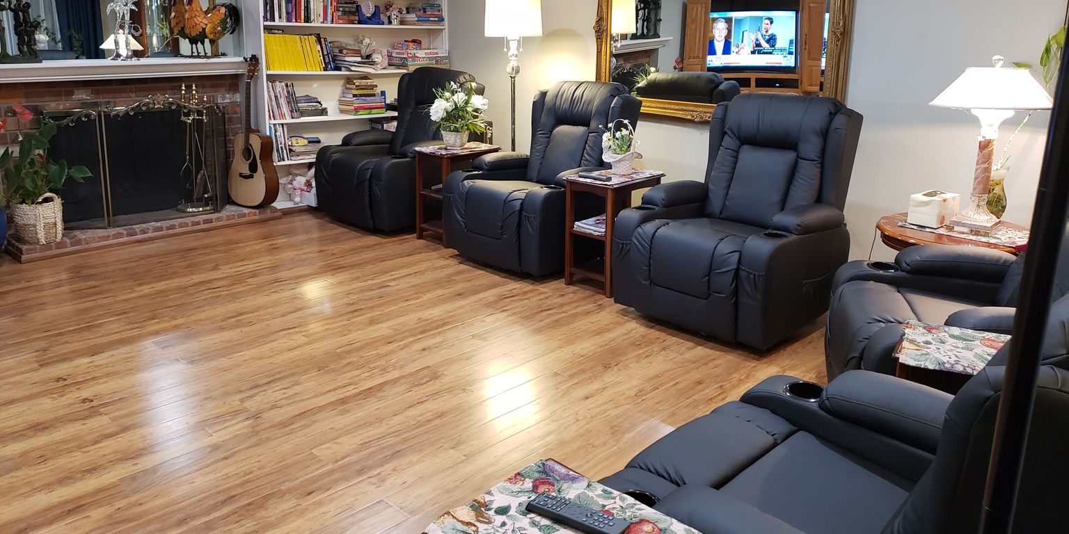A living room in Auburn adult family homes features two black leather sofas, a TV on a stand, wooden floors, a clock on the wall, large windows with curtains, and a side table with a lamp.
