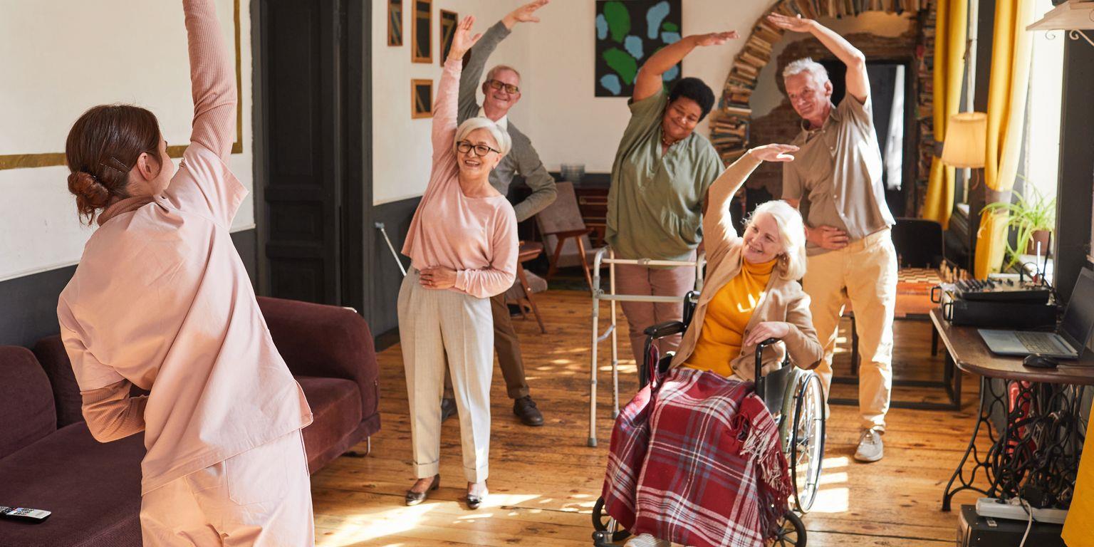 An elderly woman and a younger woman smile closely together indoors, with an orange wall and framed art in the background.