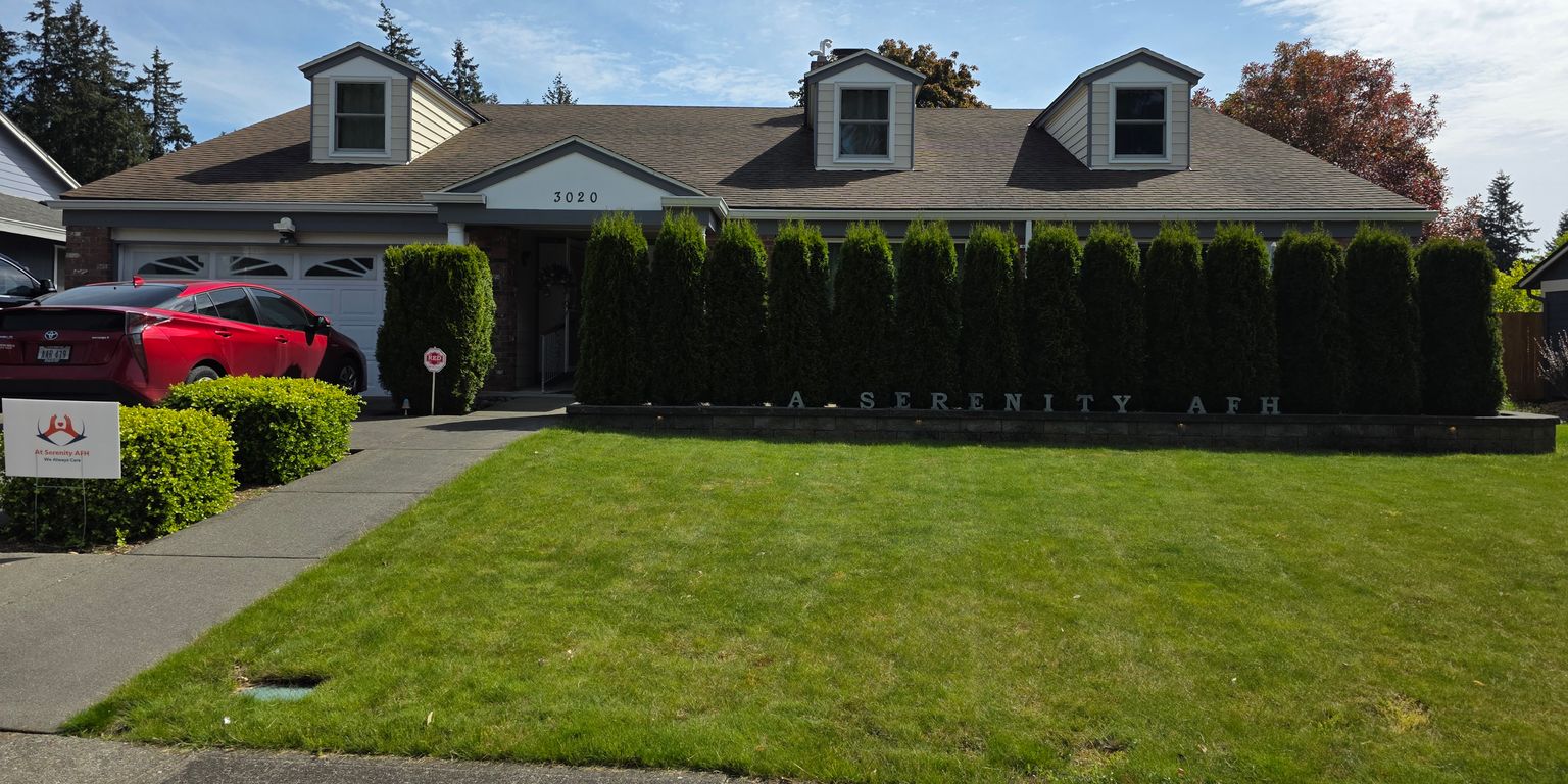 A dining room with a table, six wooden chairs, a tablecloth with a floral pattern, a vase of purple tulips, wall art, a large plant, and two ceiling lights.